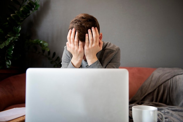 Er fühlt sich müde Frustrierter junger Mann, der erschöpft aussieht und sein Gesicht mit den Händen bedeckt, während er an seinem Laptop sitzt, seinem Arbeitsplatz zu Hause