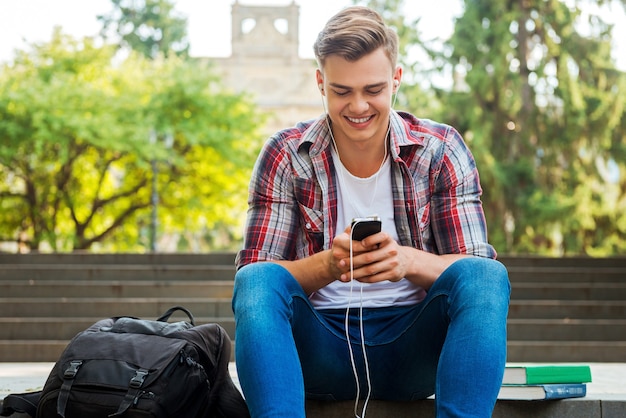 Er genießt seine Lieblingsmusik. Glücklicher männlicher Student, der MP3-Player hört und lächelt, während er an der Außentreppe sitzt, mit Büchern und Rucksack, die in seiner Nähe liegen?