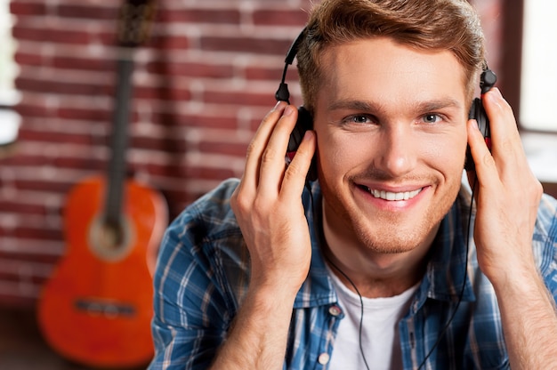 Er genießt seine Lieblingsmusik. Hübscher junger Mann, der seine Kopfhörer justiert und lächelt, während er akustische Gitarre im Hintergrund legt