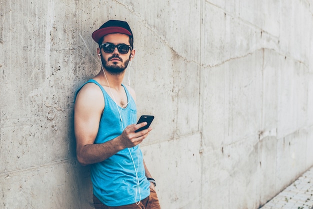 Er genießt seinen Streetstyle. Schöner junger Mann mit Kopfhörern, der ein Smartphone hält und wegschaut, während er im Freien steht und sich an die Betonwand lehnt