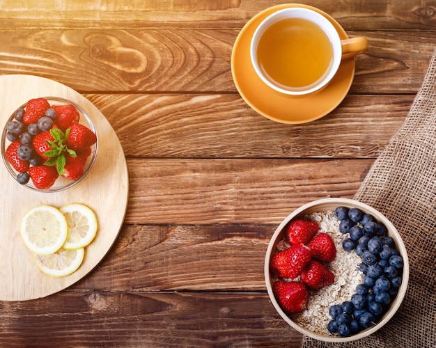 Erdbeeren, Blaubeeren und Haferflocken in der Schüssel, Tasse Tee und Zitrone auf Holzhintergrund