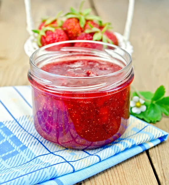 Erdbeermarmelade in einem Glas, Erdbeeren in einem Weidenkorb, Erdbeerblätter und Blumen, Serviette auf Holzbrett