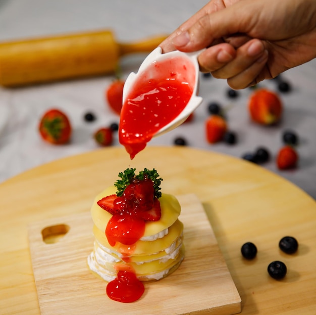 Erdbeersauce auf köstlichen Crêpe-Kuchen gießen, dekoriert mit frischen Erdbeeren auf einem Holzteller, zum Geburtstag, Valentinstag und Feiern