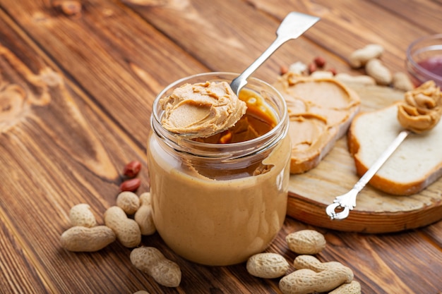 Erdnussbutter im Löffel nahe cremiger Erdnusspaste im offenen Glas, Scheibe Erdnussbutterbrot, Toast