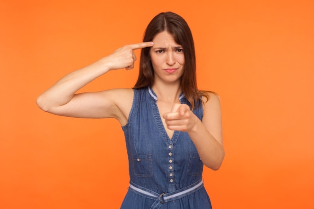 Eres idiota Mujer morena positiva con vestido de mezclilla sosteniendo el dedo cerca de la sien de la cabeza y mostrando un gesto estúpido apuntando a la cámara con una expresión burlona en un estudio aislado en un fondo naranja