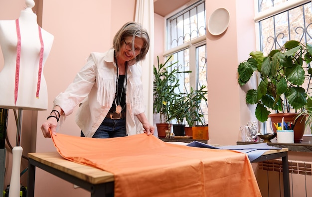 Erfahrene ältere Modedesignerin schneidert einen orangefarbenen Stoff auf einem Holztisch und kreiert ein neues Kleidungsstück im Schneideratelier