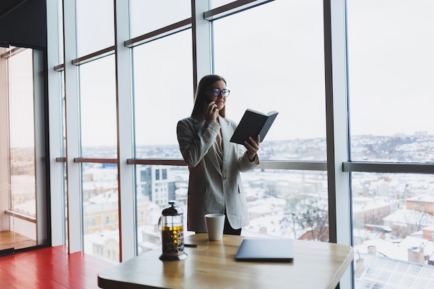 Erfahrene Geschäftsfrau, die ein Telefon hält, während sie in einem modernen Bürointerieur in der Nähe eines großen Fensters mit Blick auf das Stadtbild steht Weibliche Führungskraft sieht nach einem erfolgreichen Meeting zufrieden aus