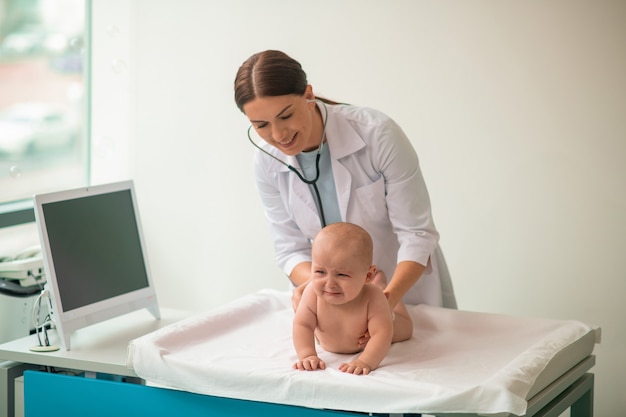 Erfahrener Arzt, der ein verärgertes Baby mit einem sterilen Stethoskop untersucht