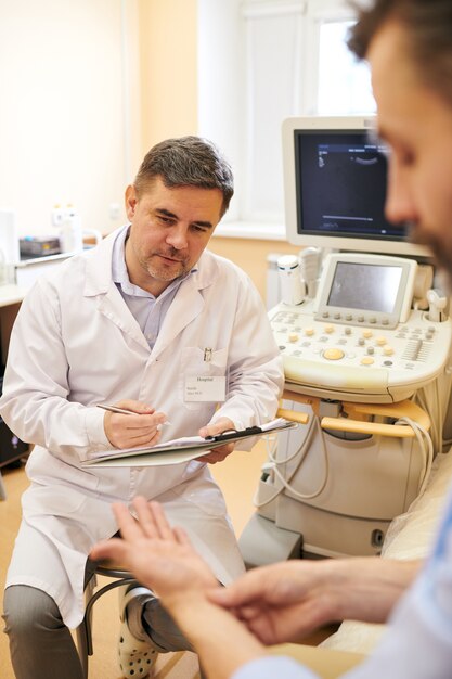 Erfahrener reifer Arzt im Laborkittel mit Zwischenablage, der Patienten zuhört, beschwert sich im Sonographieraum