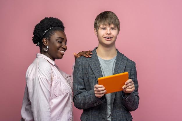 Foto erfolg. dunkelhäutige lächelnde frau seitlich zur kamera, die die schulter eines fröhlichen mannes mit down-syndrom berührt, der eine tablette auf hellem hintergrund hält