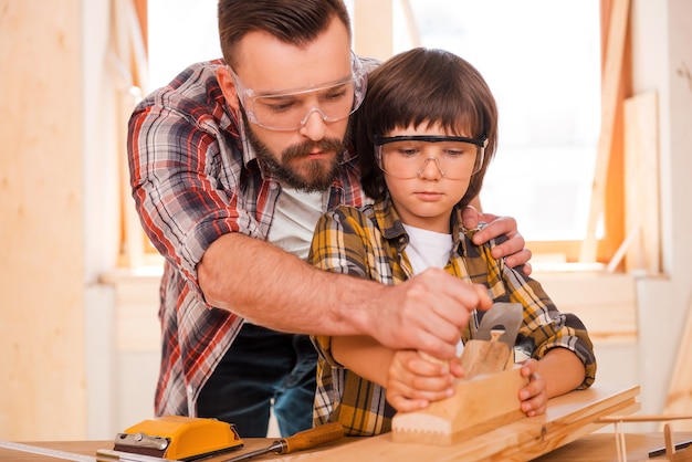 Erfolg ist eine erlernbare Fähigkeit. Konzentrierter junger männlicher Tischler, der seinem Sohn beibringt, mit Holz in seiner Werkstatt zu arbeiten