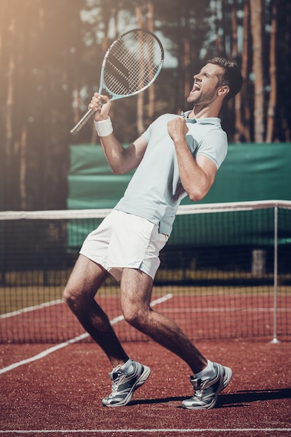 Erfolge feiern. Seitenansicht eines glücklichen jungen Mannes im Poloshirt, der Tennisschläger hält und gestikuliert, während er auf dem Tennisplatz steht