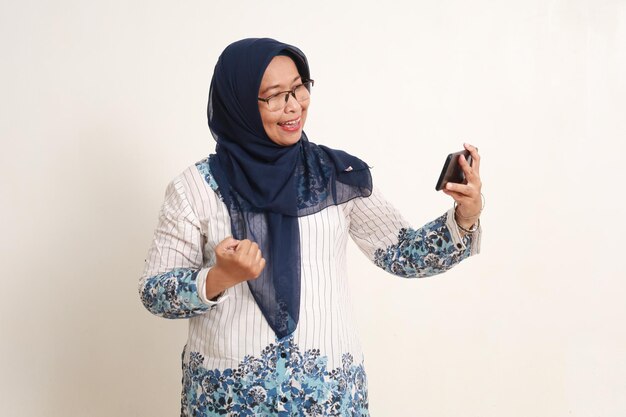 Erfolgreiche asiatische ältere muslimische Frau beobachtet Video-Streaming auf seinem Handy, während sie die Hand zusammenschlägt