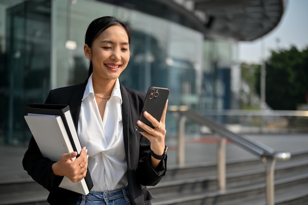 Erfolgreiche asiatische Geschäftsfrau, die ihr Smartphone benutzt, während sie die Straße entlang geht