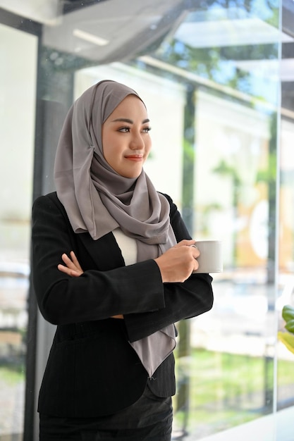 Erfolgreiche asiatische muslimische Geschäftsfrau, die aus dem Fenster schaut und vom Erfolg träumt