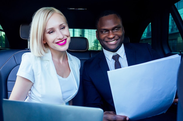 Erfolgreiche blonde Geschäftsfrau Unternehmer Arbeitgeber mit Make-up in weißem Business-Kleid mit einem gutaussehenden afroamerikanischen Chef-Mann-Industriellen in schwarzer stylischer Anzugjacke, die im Auto mit Laptop arbeitet