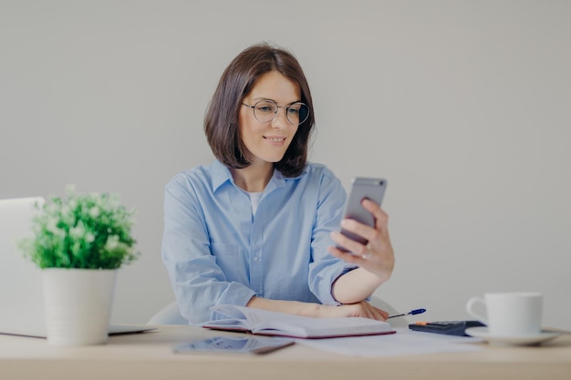 Erfolgreiche brünette Freiberuflerin erhält gute Nachrichten auf dem Smartphone, während sie am Laptop arbeitet, Beiträge auf der Website liest, Notizen im Tagebuch macht, starken Kaffee trinkt, einige Berechnungen anstellt