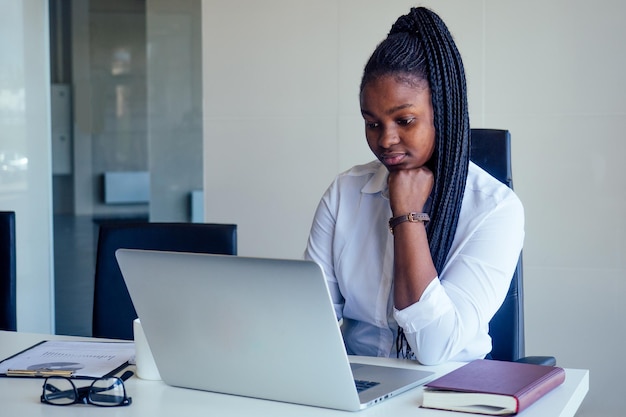 Erfolgreiche, charmante Afroamerikanerin mit Afro-Zöpfen, weißem Hemd und grauem Bleistiftrock, die in einem modernen Büro mit Panoramafenster posiert