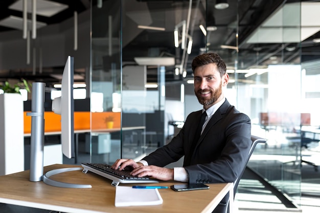 Erfolgreiche Geschäftsfrau blickt in die Kamera und lächelt glücklicher Chef, der in einem modernen Büro am Computer arbeitet