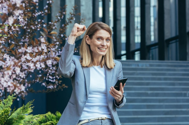 Erfolgreiche Geschäftsfrau in einem Business-Anzug außerhalb des Büros liest gute Nachrichten vom Telefon, lächelt und freut sich über den Sieg, eine Geste des Triumphs und des Sieges