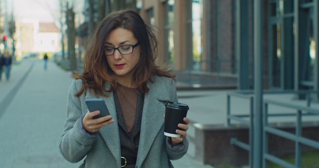 Erfolgreiche Geschäftsfrau mit Brille, die mit Kaffee geht und Smartphone im Geschäftsviertel benutzt.