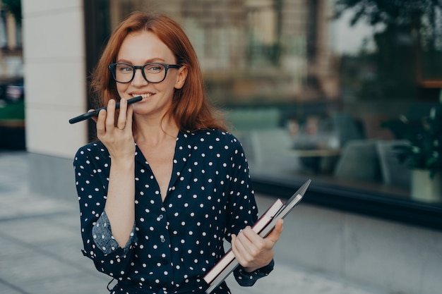Erfolgreiche Geschäftsfrau mit roten Haaren zeichnet Sprachnachrichten auf und hält Smartphone in der Nähe des Mundes