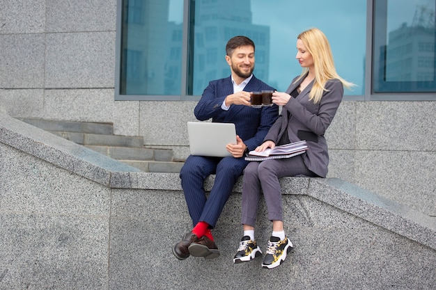 Erfolgreiche Geschäftsleute sitzen auf der Treppe eines Geschäftsgebäudes mit Dokumenten und einem Laptop in der Hand, trinken Kaffee und besprechen einen Geschäftsarbeitsplan