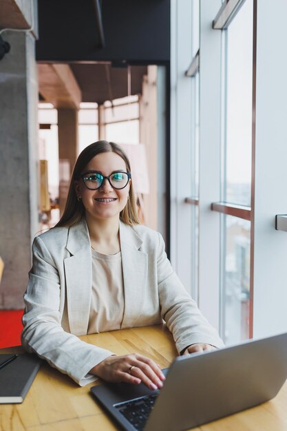 Erfolgreiche glückliche junge Frau in Jacke lächelt und arbeitet am Laptop in einem modernen Büro mit großen Fenstern Fernarbeit