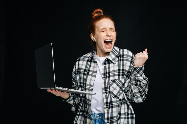 Erfolgreiche, glückliche, schockierte junge frau mit offenem mund und geschlossenen augen, die einen laptop auf isoliertem schwarzem hintergrund hält hübsches damenmodell mit roten haaren, das emotional gesichtsausdrücke zeigt