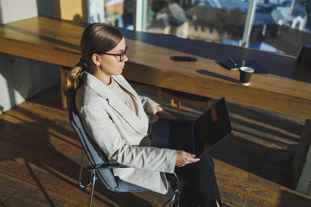 Erfolgreiche junge Geschäftsfrau in Gläsern mit Laptop im Büro Geschäftsfrau, die E-Mail-Nachricht sendet und am Laptop arbeitet Junge Geschäftsfrau, die am Arbeitsplatz sitzt