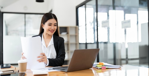 Erfolgreiche lächelnde Geschäftsfrau benutzt Laptop und Computer, während sie Papierarbeit im Büro hält