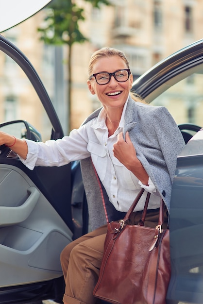 Erfolgreiche modische Geschäftsfrau mittleren Alters mit Handtasche, die aus ihrem modernen Auto aussteigt und