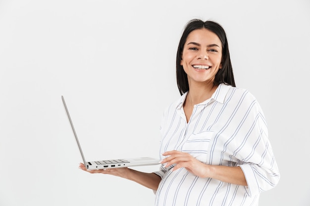 erfolgreiche schwangere Frau mit großem Bauch, der lächelt und Laptop in Händen hält, während lokalisiert über weißer Wand steht