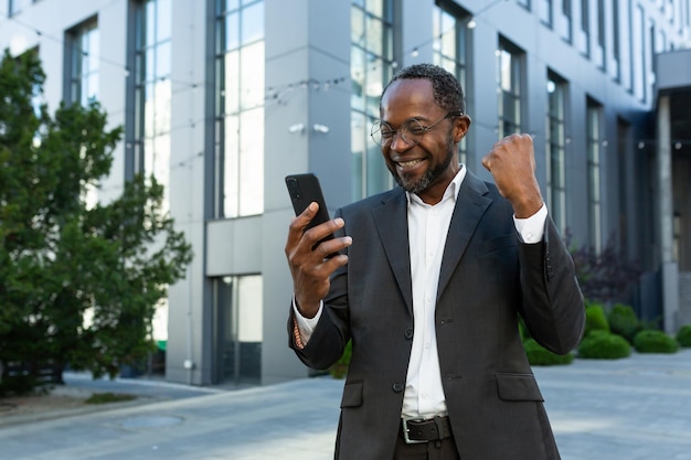 Erfolgreicher afrikanisch-amerikanischer reifer Investor mit Telefon vor dem Bürogebäude, der frohe Nachrichten liest