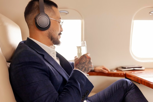 Erfolgreicher asiatischer Geschäftsmann fliegt mit einem Glas Champagner in einem Privatflugzeug und hört Musik