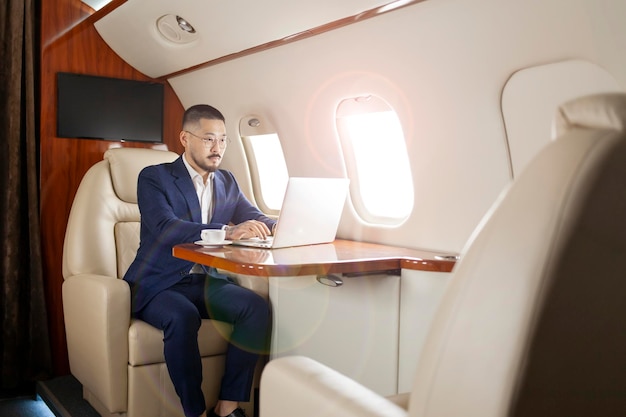 Erfolgreicher asiatischer Geschäftsmann in Anzug und Brille sitzt im Privatjet und tippt auf dem Laptop