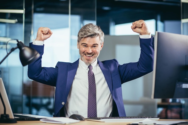 Erfolgreicher Chef arbeitet in einem modernen Büro am Computer, ein älterer Mann, grau und glücklich mit Bart, schaut auf die Kamera und lächelt, hält seine Hände hoch und feiert den Sieg