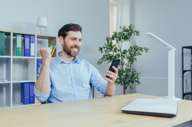 Erfolgreicher freiberuflicher Geschäftsmann arbeitet im Home Office, schaut auf den Telefonbildschirm und feiert den Sieg, hält die Hände hoch und freut sich über eine triumphale Geste