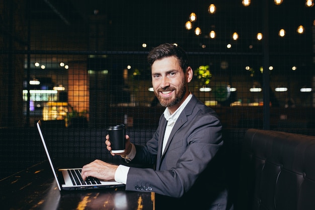 Erfolgreicher Geschäftsmann, der in der Mittagspause am Laptop arbeitet und im Hotelrestaurant sitzt, lächelt mit einer Tasse Kaffee in die Kamera