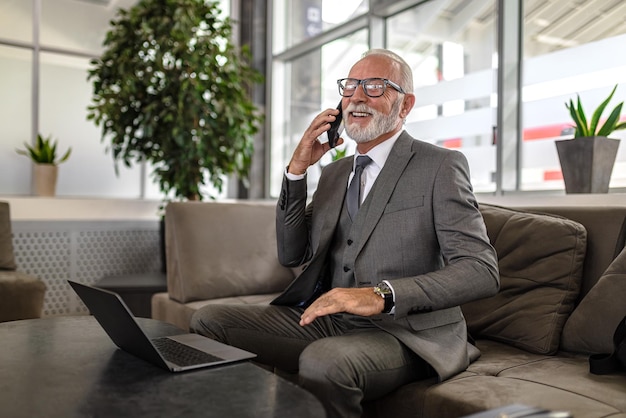 Erfolgreicher Geschäftsmann, der mit dem Smartphone telefoniert, während er im Bürosaal einen Laptop benutzt