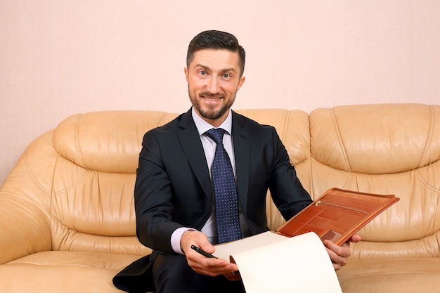 Erfolgreicher Geschäftsmann, der mit Geschäftsdokumenten lächelt, die auf einem Ledersofa sitzen