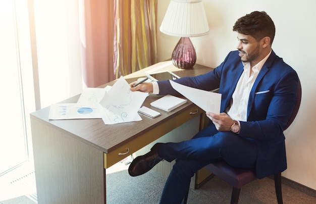 Erfolgreicher Geschäftsmann, der mit Papieren arbeitet, während er am Tisch im Hotelzimmer sitzt