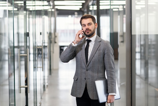 Erfolgreicher Geschäftsmann hat Telefongespräch, während er im leeren Bürokorridor steht. Männlicher professioneller Bankier im Anzug, der während der Arbeitspause auf Mobiltelefon spricht.