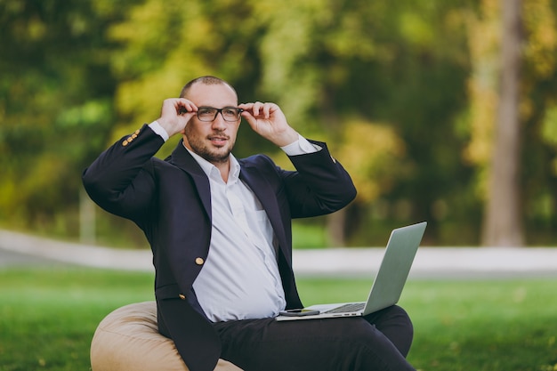 Erfolgreicher Geschäftsmann im weißen Hemd, im klassischen Anzug, in der richtigen Brille. Mann sitzt auf weichem Hocker, arbeitet an Laptop-PC im Stadtpark auf grünem Rasen im Freien in der Natur. Mobiles Büro, Geschäftskonzept.