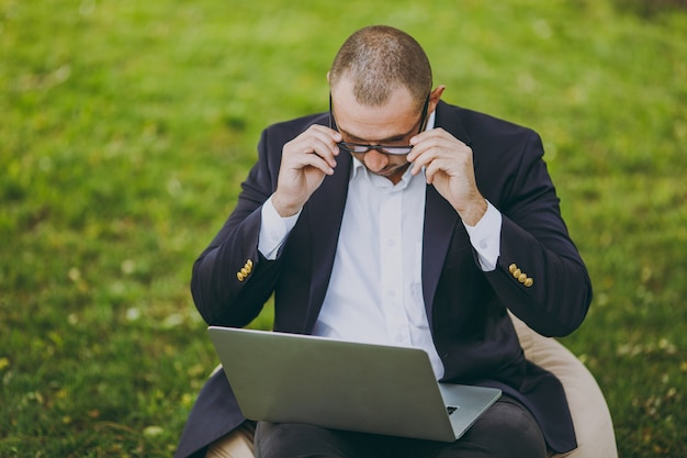 Erfolgreicher Geschäftsmann im weißen Hemd, im klassischen Anzug, in der richtigen Brille. Mann sitzt auf weichem Hocker, arbeitet an Laptop-PC im Stadtpark auf grünem Rasen im Freien in der Natur. Mobiles Büro, Geschäftskonzept.