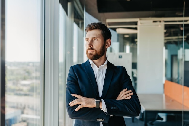 Erfolgreicher Geschäftsmann in formeller Kleidung, der mit verschränkten Armen in einem modernen Coworking-Büro steht und durch das Fenster schaut