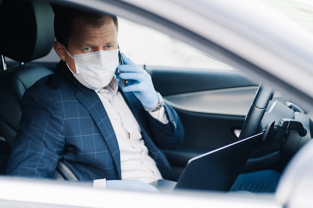 Erfolgreicher Geschäftsmann wartet auf jemanden im Auto, setzt während der Epidemie eine medizinische Maske als Schutz vor Viren auf, telefoniert, benutzt einen modernen Laptop. Covid-19, Quarantäne, Infektion, Krankheitskonzept