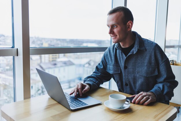 Erfolgreicher, glücklicher Geschäftsmann sitzt an einem Tisch in einem Café, hält eine Tasse Kaffee und benutzt einen Laptop