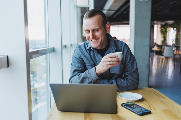 Erfolgreicher, glücklicher Geschäftsmann sitzt an einem Tisch in einem Café, hält eine Tasse Kaffee und benutzt einen Laptop