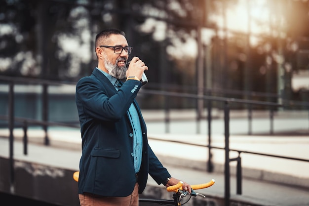 Erfolgreicher gutaussehender Geschäftsmann mittleren Alters mit Fahrrad neben ihm geht vor dem Büroviertel spazieren und trinkt einen Kaffee zum Mitnehmen.